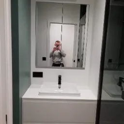 Nowoczesna łazienka z białą szafką pod umywalką, czarną armaturą i lustrem z wbudowanymi szafkami, odbicie osoby robiącej zdjęcie telefonem.