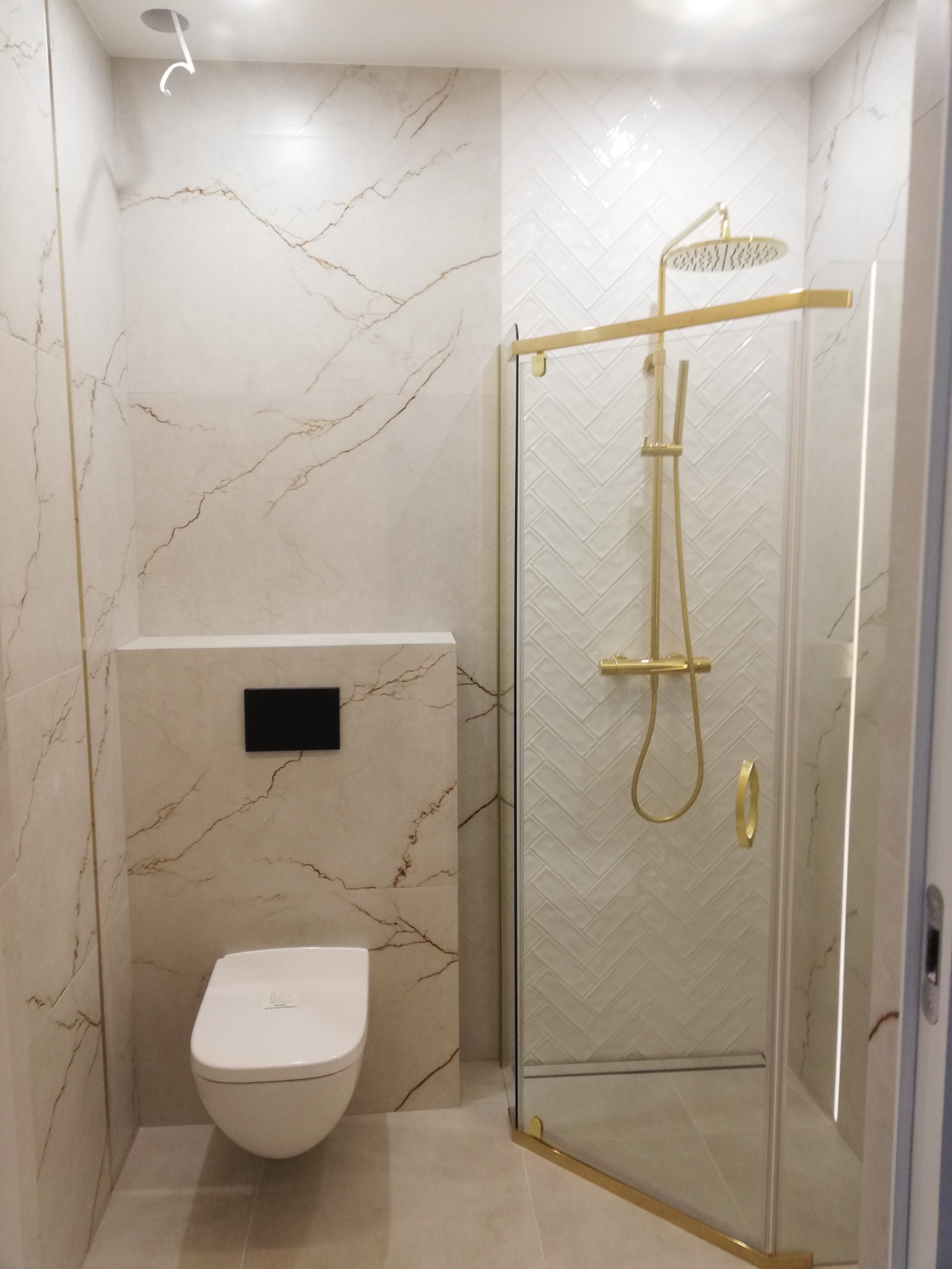 Elegancka łazienka z kabiną prysznicową ze złotymi detalami, białą armaturą i płytkami imitującymi marmur z subtelnym złotym żyłkowaniem. Widoczny podwieszany sedes.