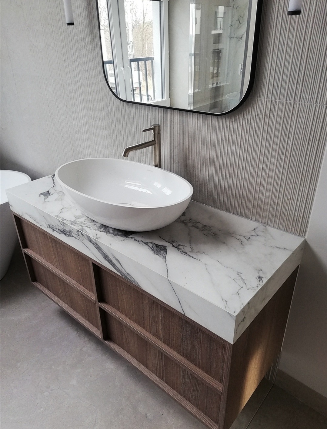 Nowoczesna łazienka: biała, owalna umywalka nablatowa na szafce z marmurowym blatem i drewnianymi frontami, lustro w czarnej ramie, minimalistyczna armatura.