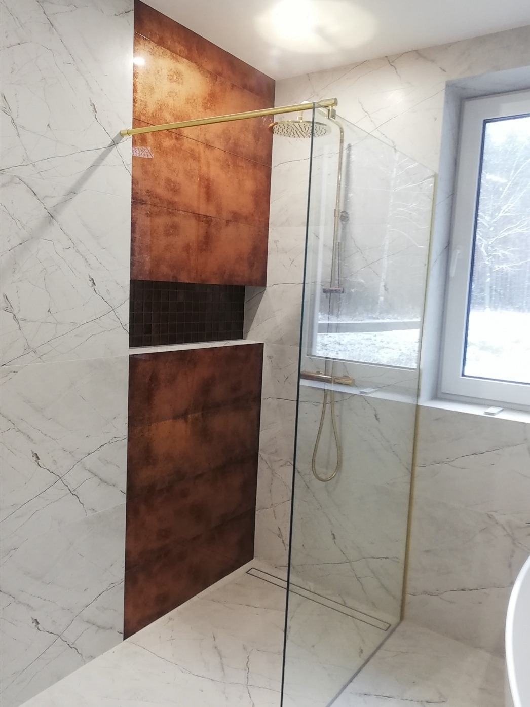 Nowoczesna łazienka z marmurowymi płytkami, kabiną prysznicową typu walk-in ze złotymi detalami i wnęką z mozaiką. Widok na okno z zimowym krajobrazem.