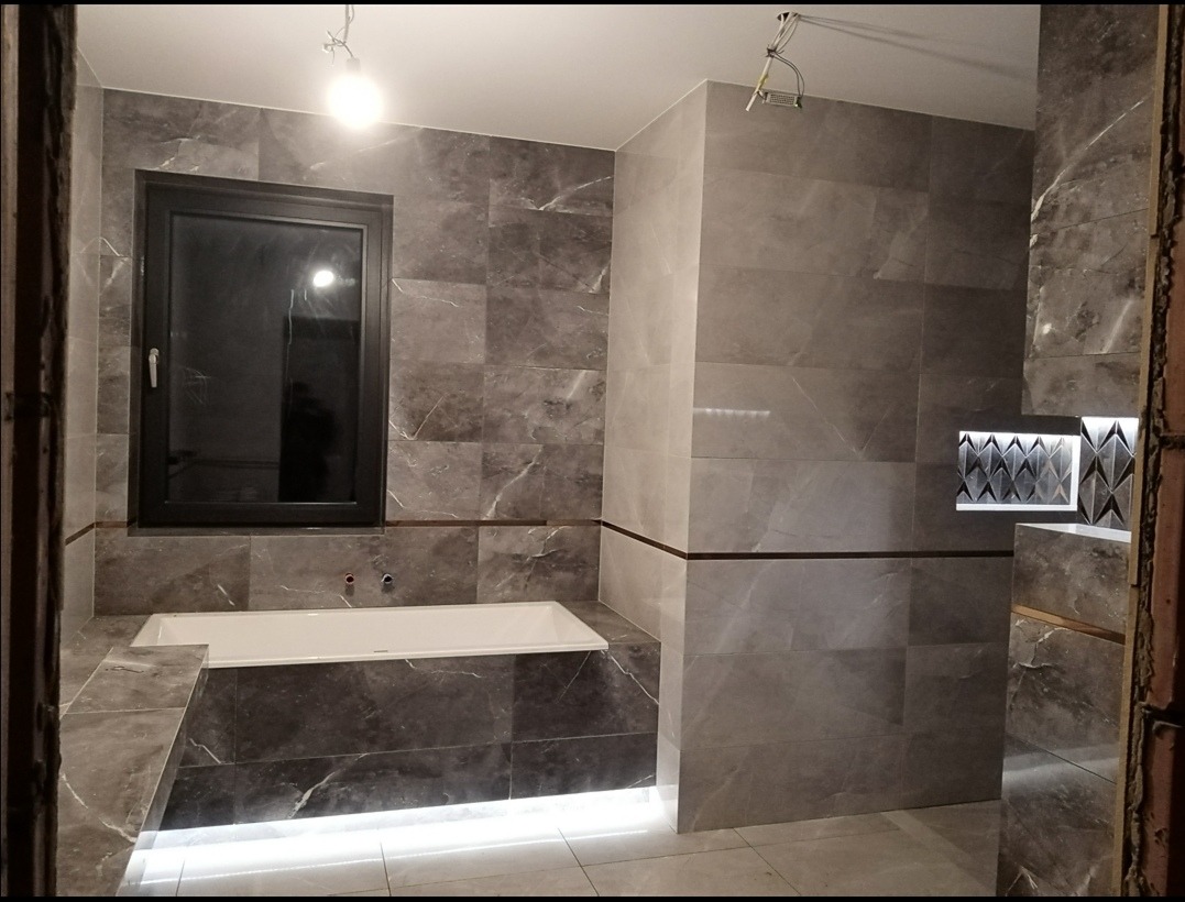 Wykończona łazienka z szarymi płytkami imitującymi marmur, podświetlana wanna, czarna rama okna i dekoracyjna wnęka z oświetleniem. Nowoczesny design.