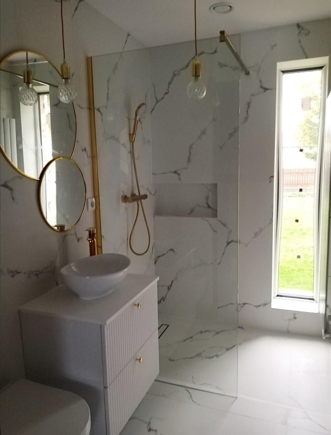 Nowoczesna łazienka z kabiną typu walk-in, białą umywalką nablatową i marmurowymi płytkami. Złote akcenty w armaturze i oświetleniu dodają elegancji.