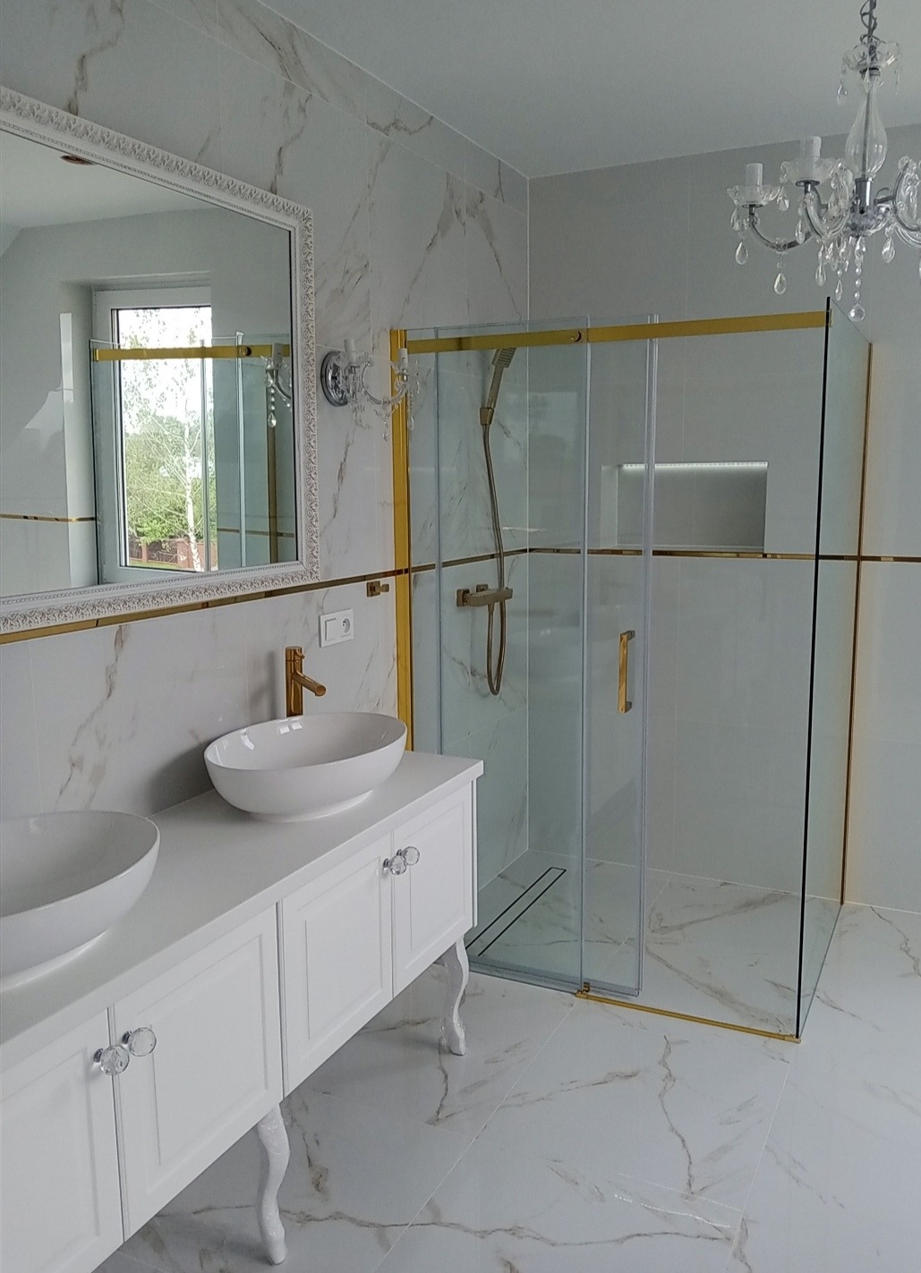 Elegancka łazienka z marmurowymi płytkami, kabiną prysznicową ze złotymi detalami i białą szafką z umywalkami. Lustro z ozdobną ramą i kryształowy żyrandol.