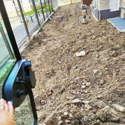 USŁUGI OGÓLNOBUDOWLANE KOPARECZKA Łukasz Luszowski - Niwelacja terenu z ubijarką stopową, widok z kabiny maszyny, przygotowany teren pod budowę, ogrodzenie z siatki, budynek w tle.