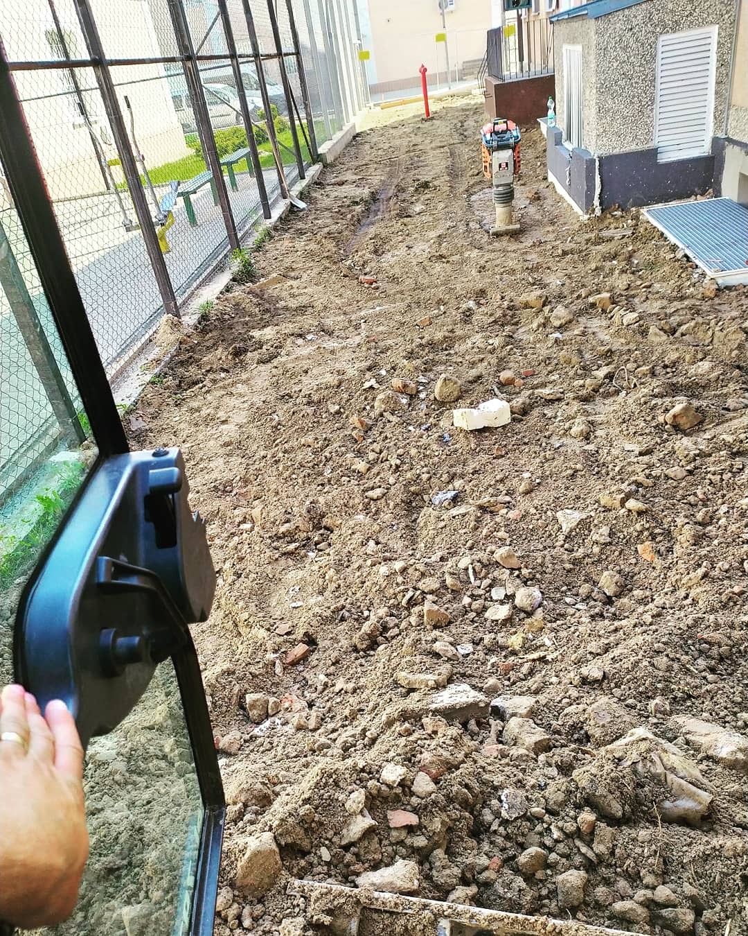 Niwelacja terenu z ubijarką stopową, widok z kabiny maszyny, przygotowany teren pod budowę, ogrodzenie z siatki, budynek w tle.