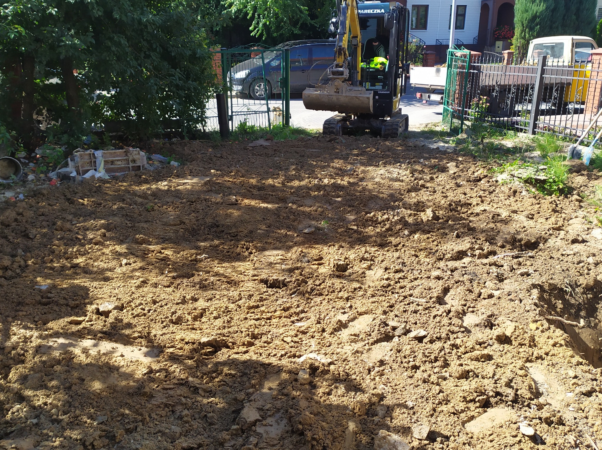 Żółta mini koparka Yanmar na wyrównanym terenie budowy, widoczne ogrodzenie i zabudowa jednorodzinna w tle.