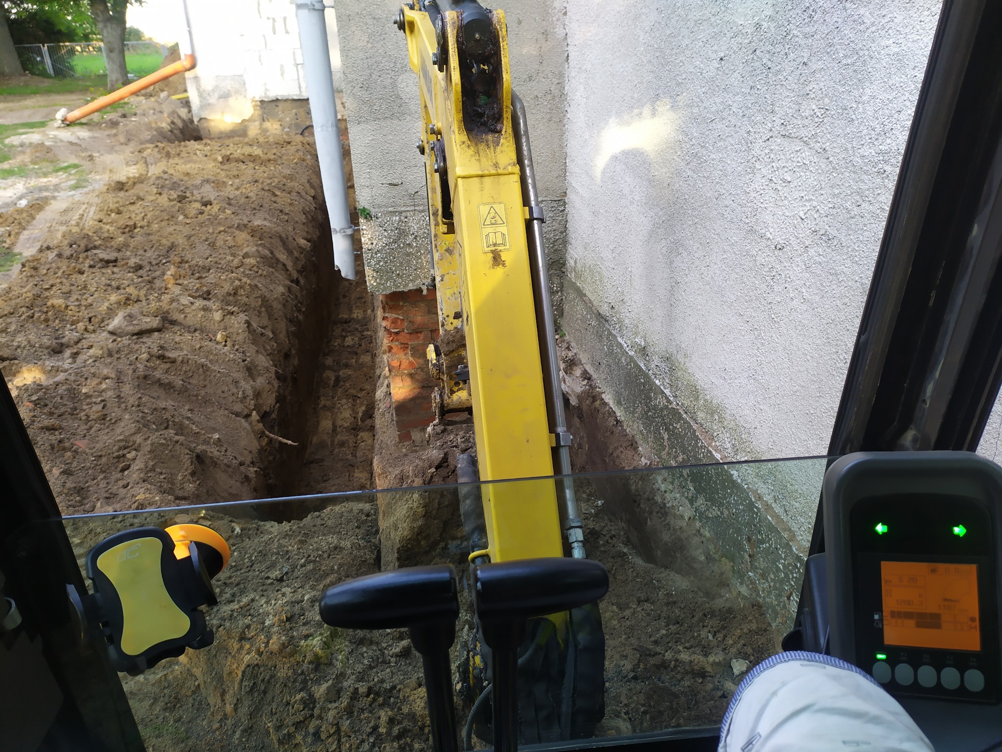 Widok z kabiny koparki na wąski wykop w ziemi przy ścianie budynku z widoczną ceglaną podmurówką.