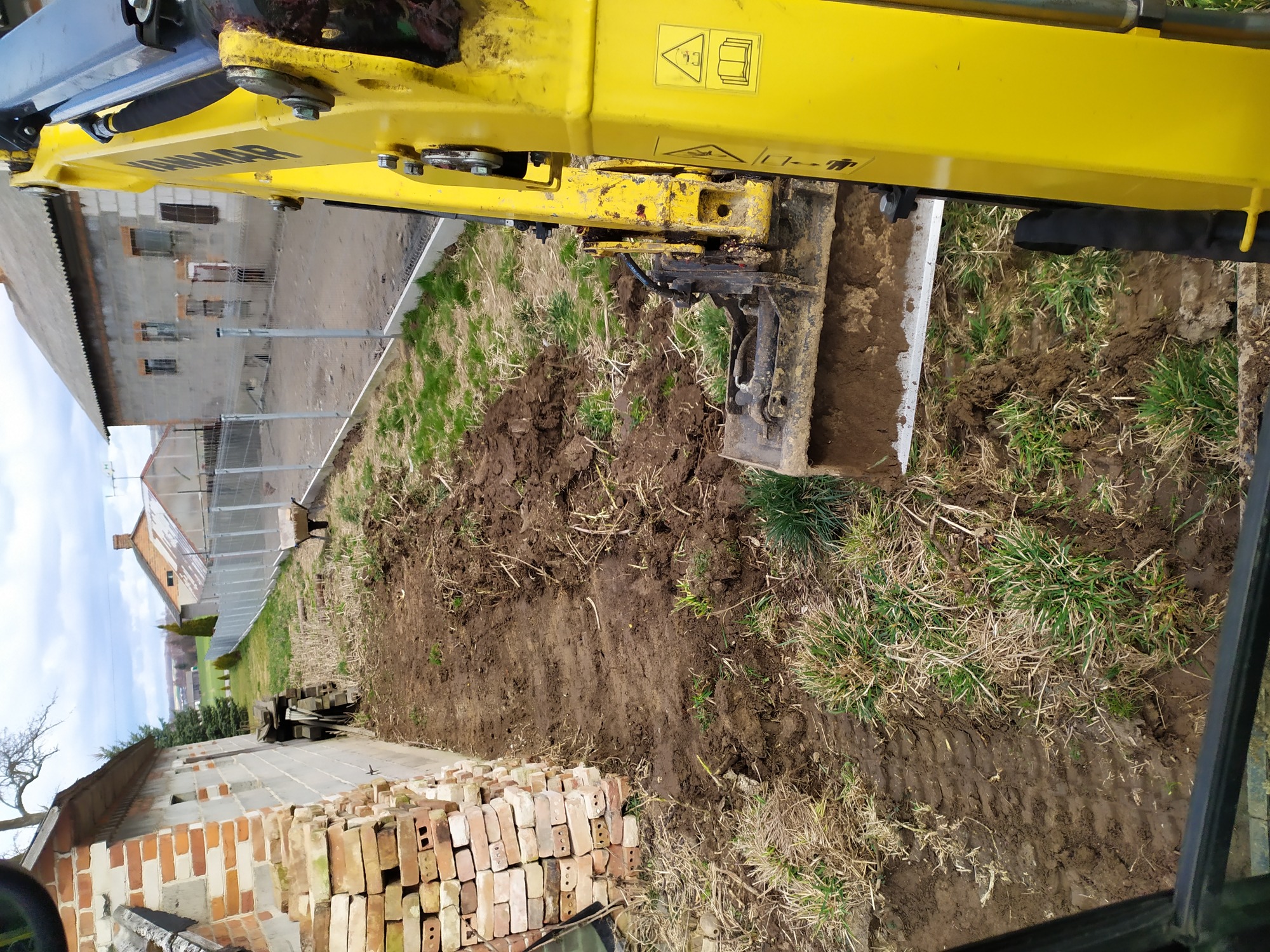 Żółta koparka Yanmar wyrównuje teren, widoczne ślady gąsienic na ubitej ziemi, w tle budynek gospodarczy i sterta cegieł.