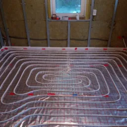 Montaż ogrzewania podłogowego w poddaszu, widoczne białe rury na folii z czerwoną siatką, ocieplenie wełną mineralną na skośnych ścianach poddasza, okno w tle.