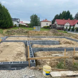 Fundamenty domu jednorodzinnego w trakcie budowy. Widoczne bloczki fundamentowe i wykop. W tle zabudowa mieszkalna i zieleń.