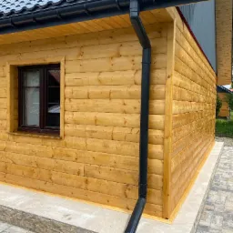 Drewniany domek z brązowym oknem i czarną rynną. Elewacja z jasnego drewna, widoczne łączenia desek. Ujęcie z boku, fragment chodnika z kostki.