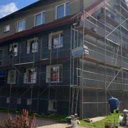 B.O.M.M.N. - Wielopiętrowy budynek w trakcie ocieplania, pokryty szarym styropianem, z widocznymi rusztowaniami i pracownikami. Częściowo ocieplona elewacja, worki z materiałami budowlanymi.