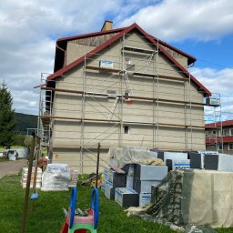 B.O.M.M.N. - Dom w trakcie ocieplania elewacji, widoczne rusztowania, materiały izolacyjne i częściowo otynkowana ściana. W tle zielony krajobraz i błękitne niebo.