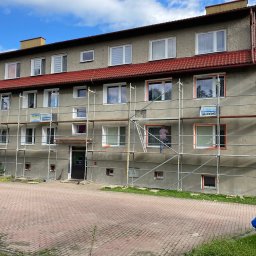 B.O.M.M.N. - Budynek wielorodzinny w trakcie ocieplania elewacji, widoczne rusztowania i pracownik przy oknie. Częściowo ocieplona ściana z zamontowanymi oknami. Czerwona dachówka.