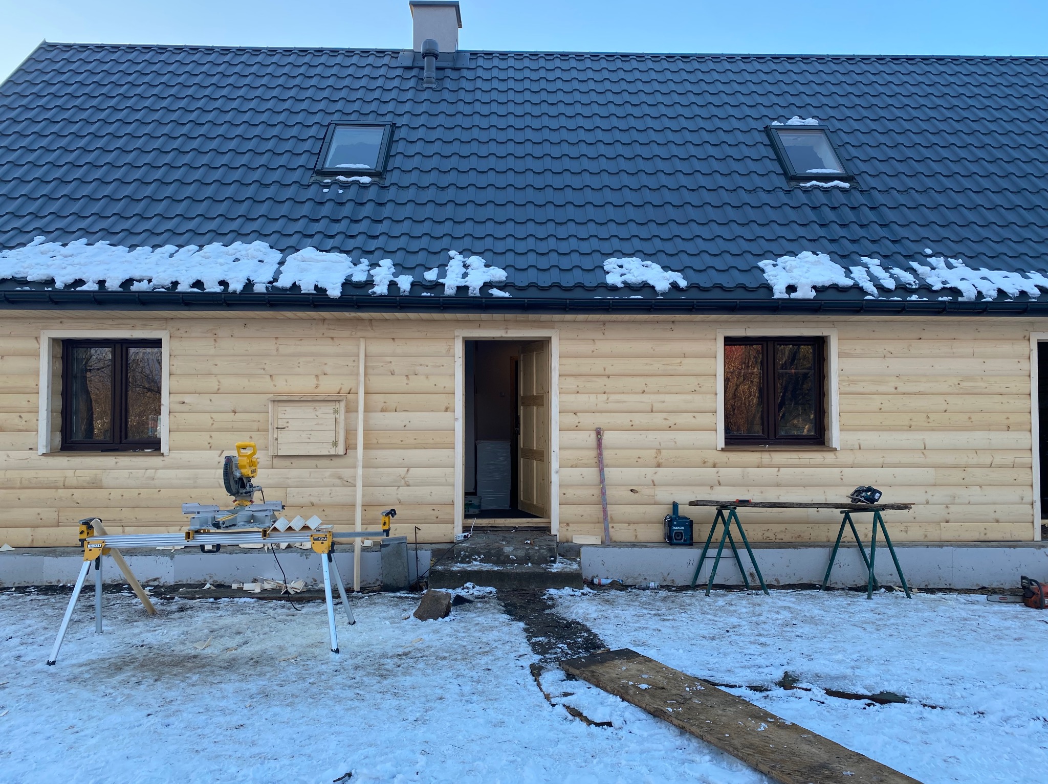 Budowa domu z drewnianych bali w zimowej scenerii, widoczna piła stołowa na zewnątrz, częściowo ośnieżony dach z oknami dachowymi, oraz surowe wykończenie elewacji.
