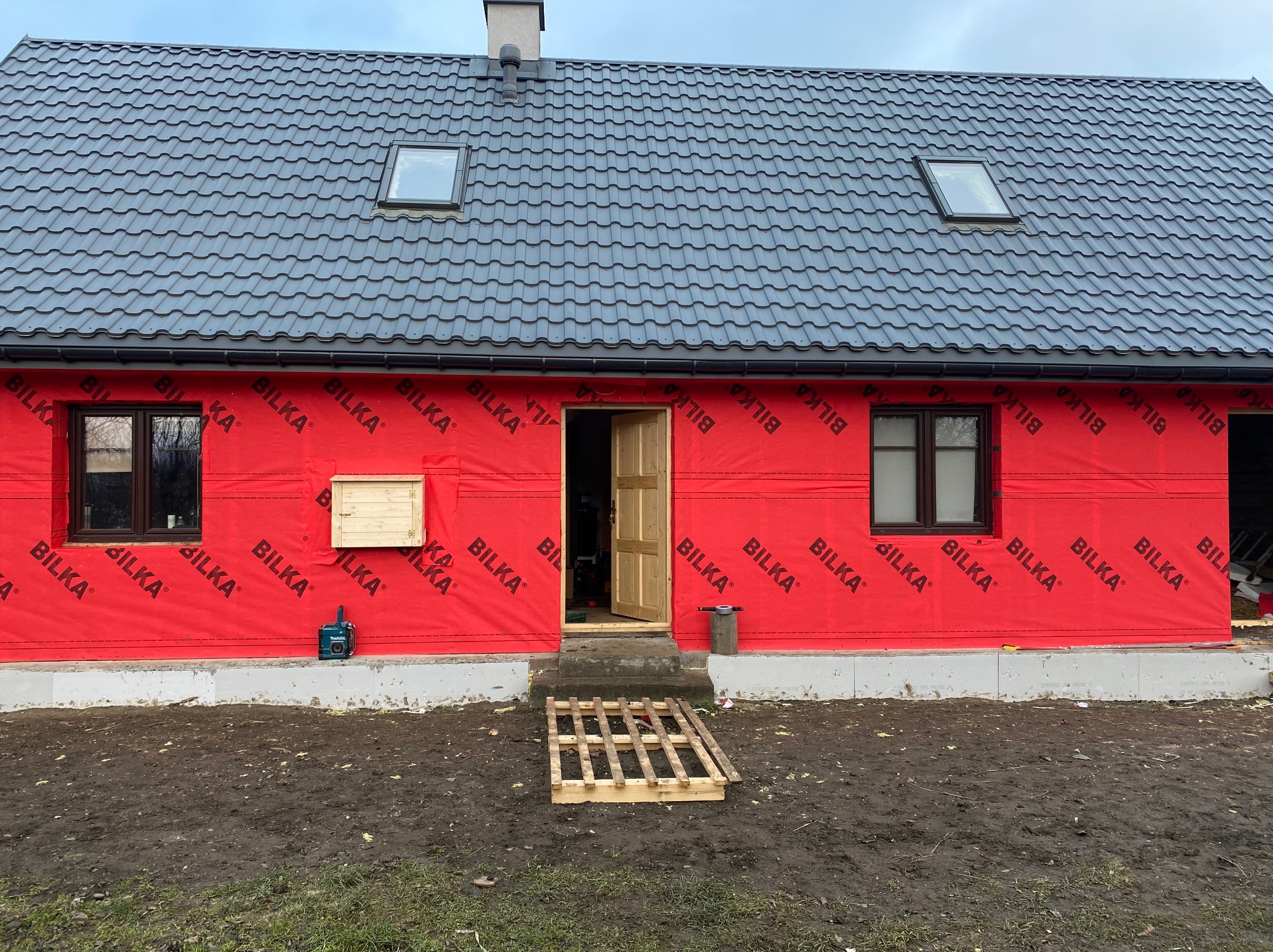 Dom w trakcie ocieplania, zabezpieczony czerwoną folią izolacyjną z widocznym logo producenta, z otwartymi drewnianymi drzwiami wejściowymi i szarym dachem z oknami połaciowymi, na nierównym...