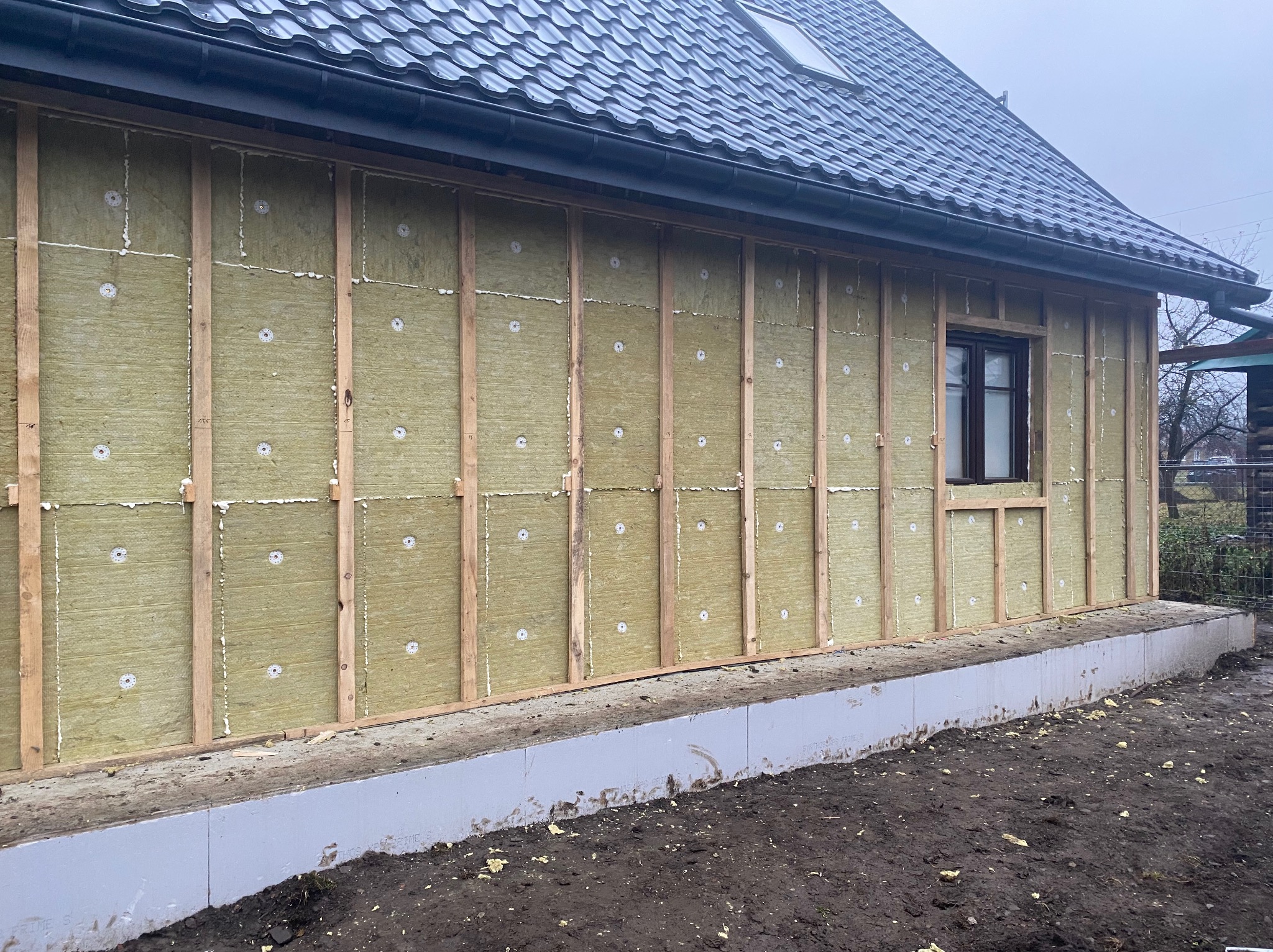 Ściana domu w trakcie ocieplania wełną mineralną, widoczne drewniane ramy konstrukcyjne i kołki mocujące izolację, szara elewacja fundamentu, dach pokryty ciemną dachówką.