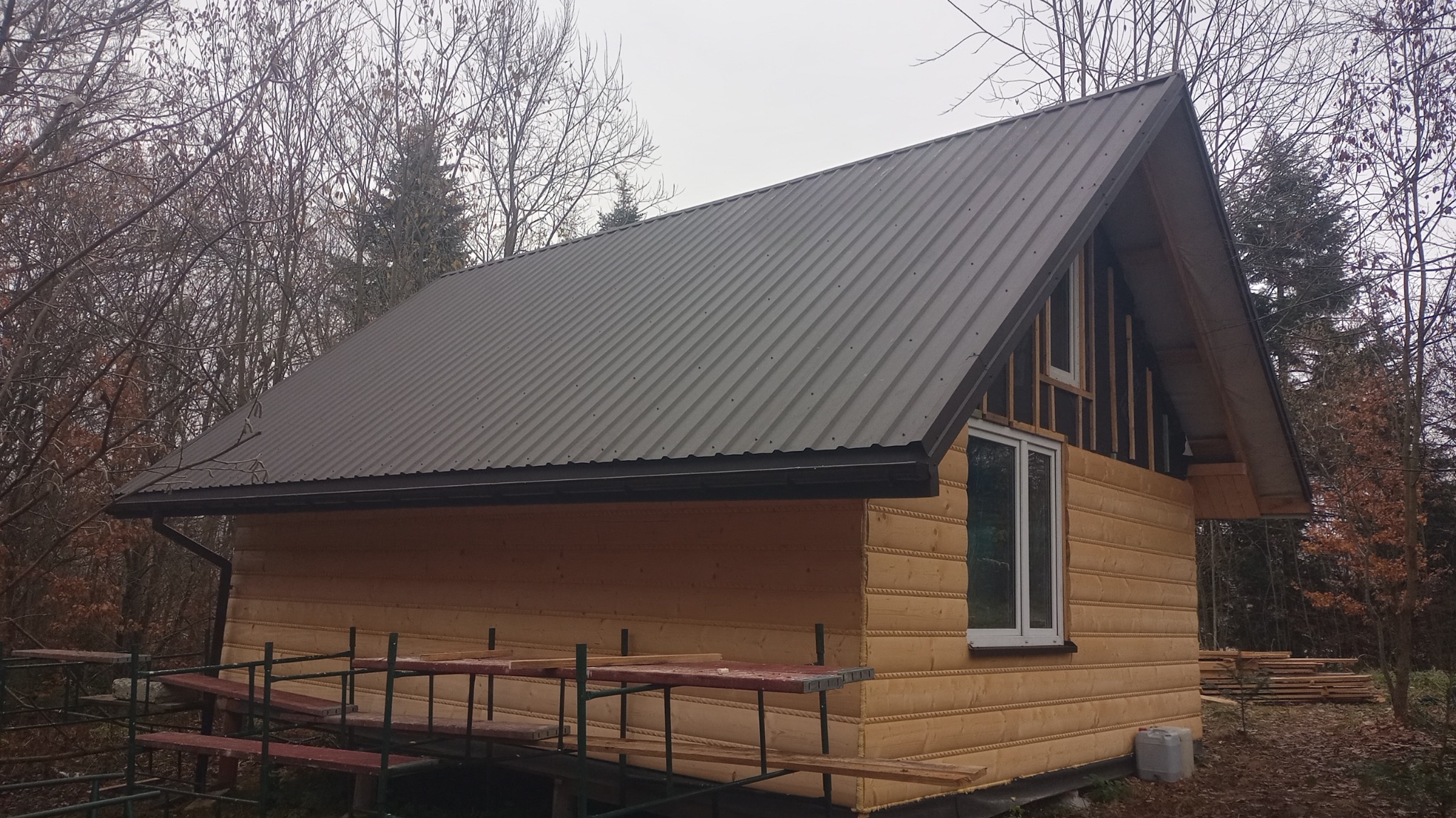 Drewniany dom w trakcie budowy z zamontowaną blachodachówką i częściowo ocieploną ścianą szczytową, widoczne rusztowanie i składowane deski.