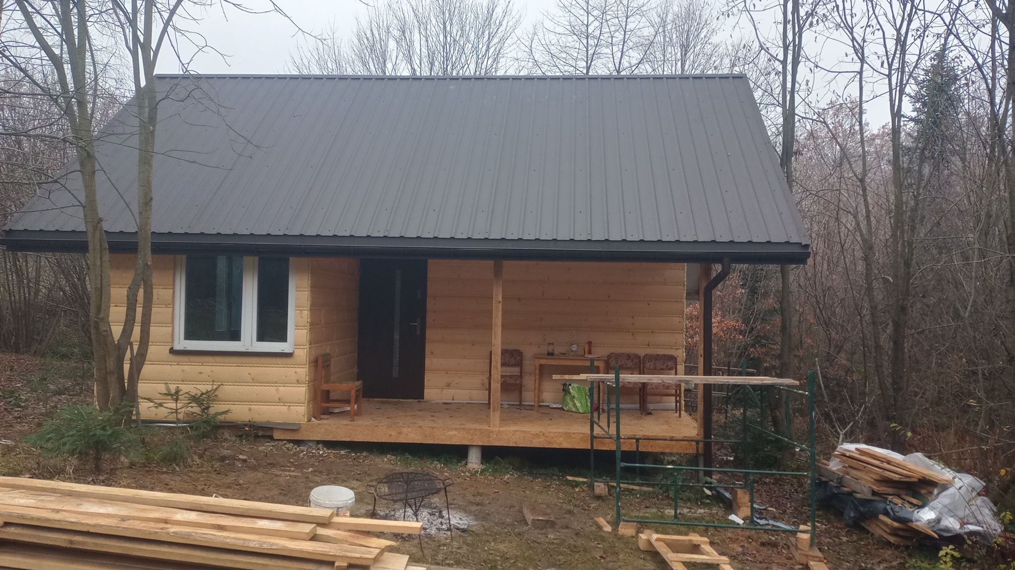 Nieduży, drewniany dom z ciemnoszarym, metalowym dachem, częściowo wykończony, z widocznym podestem wejściowym i stosem desek obok, na tle jesiennego lasu.