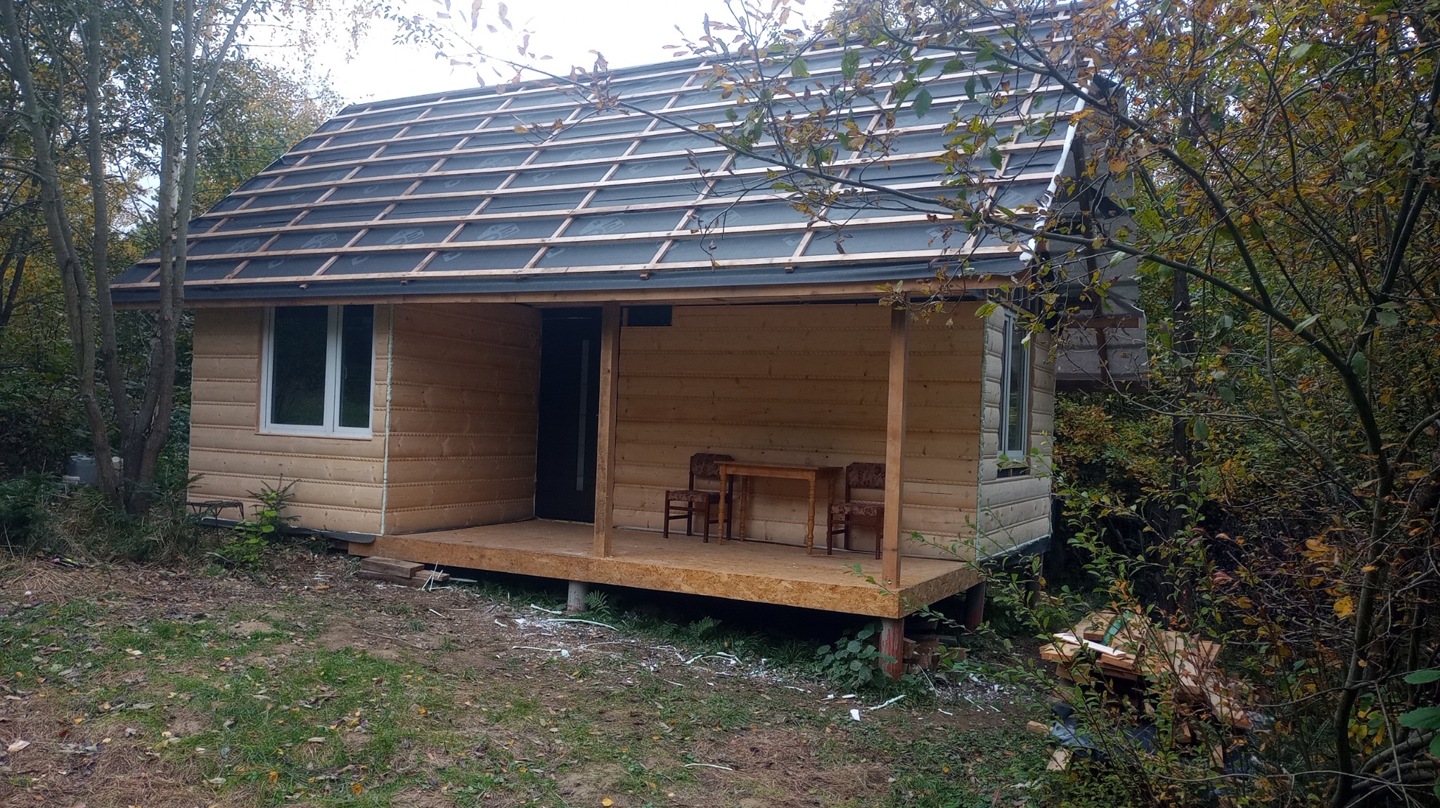 Niedokończony drewniany dom z widoczną konstrukcją dachu i deskami elewacyjnymi w otoczeniu jesiennego lasu, z małym tarasem, na którym stoją dwa krzesła i stół.