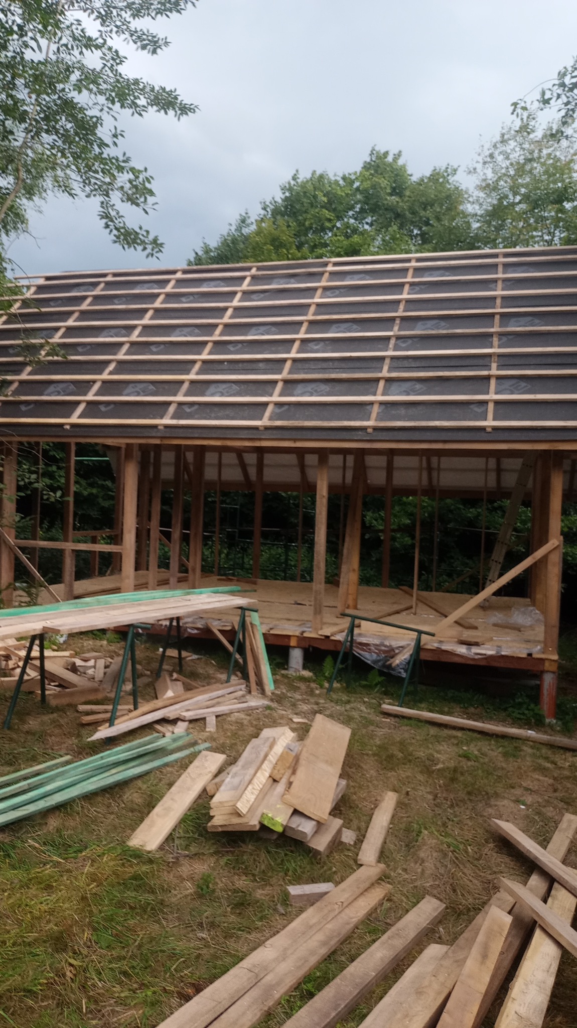 Konstrukcja drewniana domu w trakcie budowy z widocznym szkieletem i dachem pokrytym membraną, deski przygotowane do dalszych prac na trawie.