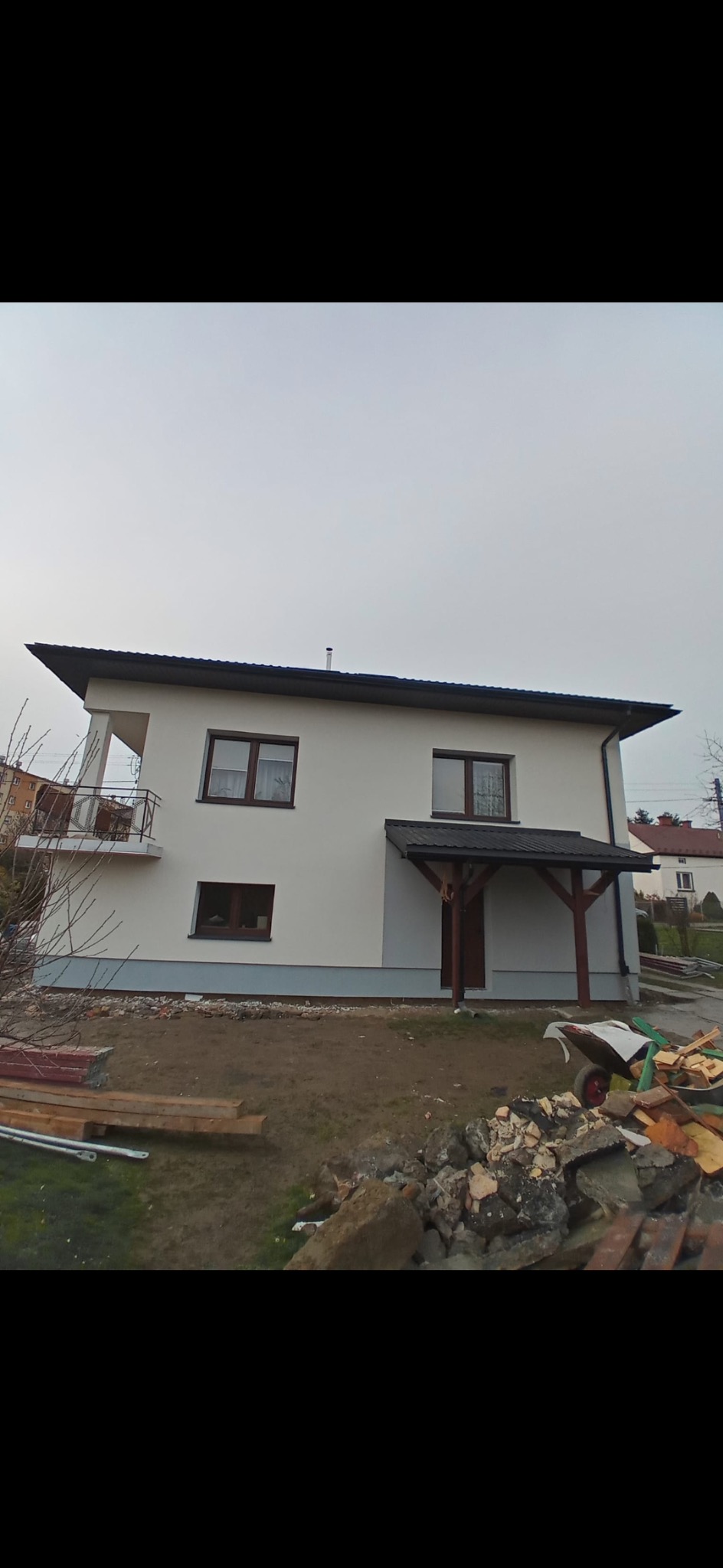 Dwukondygnacyjny dom jednorodzinny z białymi ścianami i ciemnobrązowymi oknami, zadaszonym wejściem i balkonem, widok z zewnątrz na tle pochmurnego nieba, teren wokół domu w trakcie zagospodarowania.