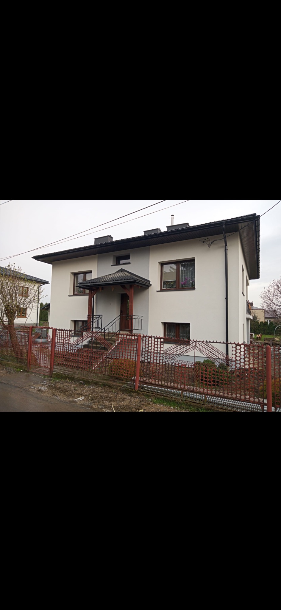 Dwukondygnacyjny dom murowany z jasną elewacją, ciemnym dachem i drewnianym zadaszeniem nad wejściem, otoczony czerwonym, ażurowym ogrodzeniem.