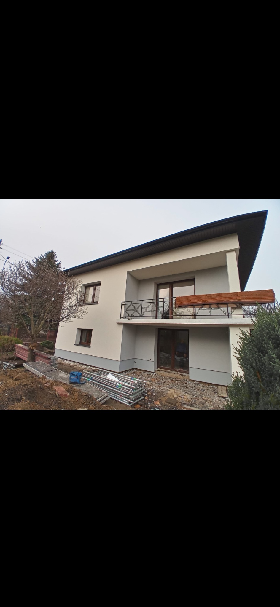 Nowoczesny dom murowany z balkonem i elementami drewnianymi, widoczne rusztowania i teren w trakcie zagospodarowywania.