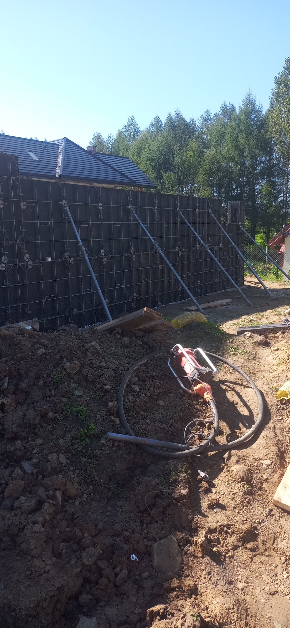 Szare szalunki fundamentowe wzmocnione metalowymi stężeniami, widoczny wibrator do betonu leżący na ziemi, w tle fragment dachu domu i drzewa.