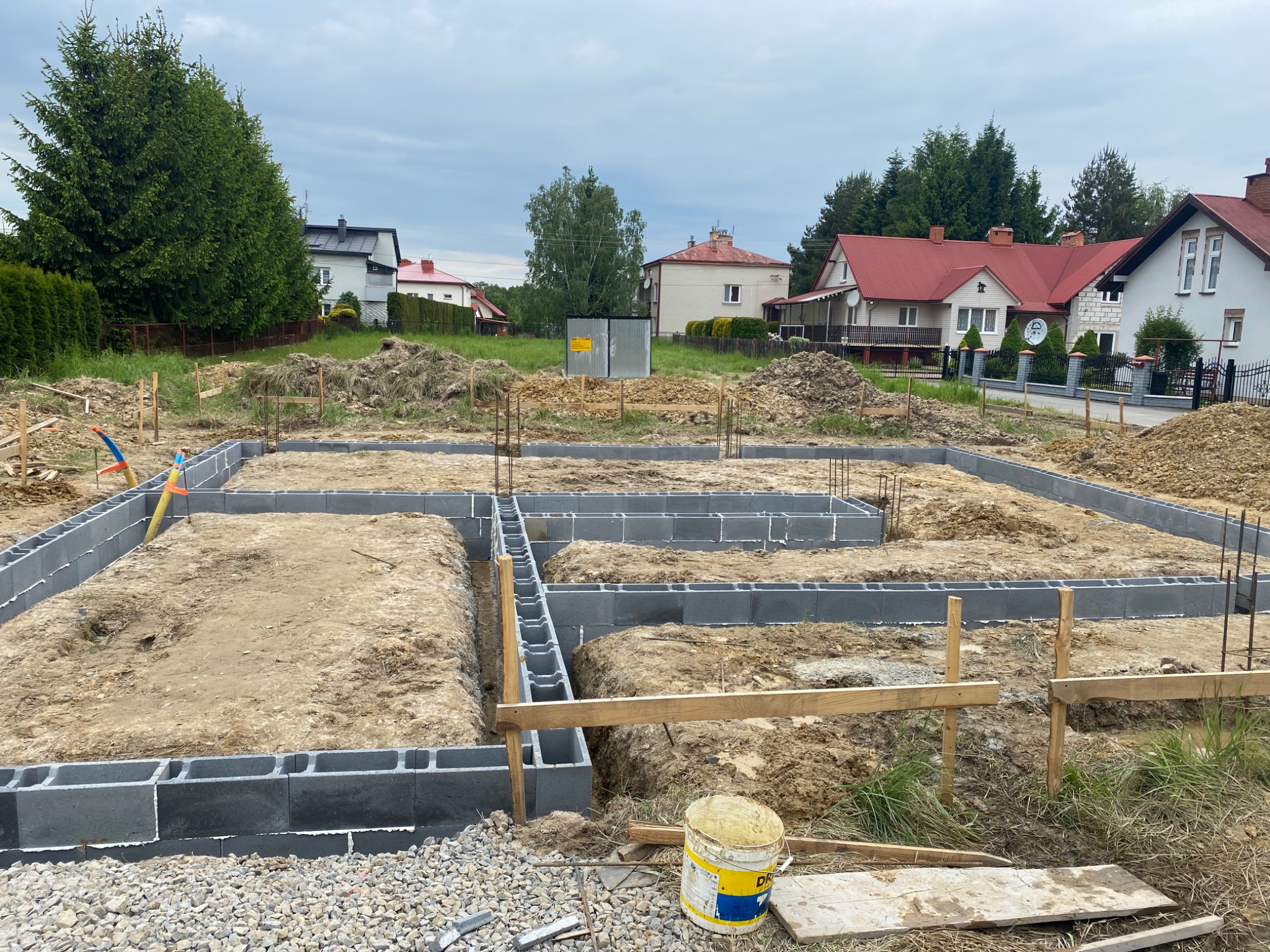Fundamenty domu jednorodzinnego w trakcie budowy. Widoczne bloczki fundamentowe i wykop. W tle zabudowa mieszkalna i zieleń.