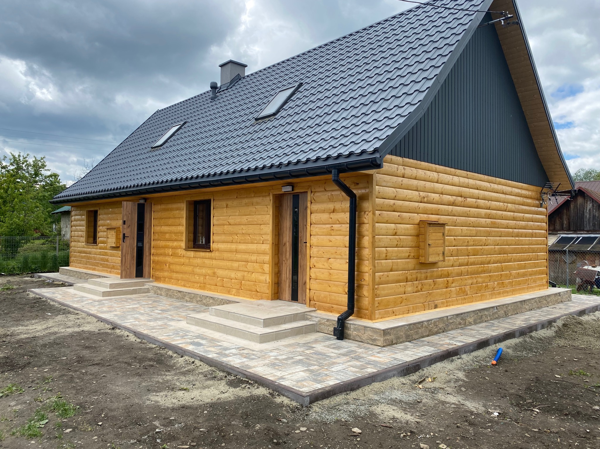 Nowoczesny dom szkieletowy z drewnianą elewacją i szarym dachem dwuspadowym. Widoczne okna dachowe i wykończone wejście z kostki brukowej. Pochmurne niebo w tle.