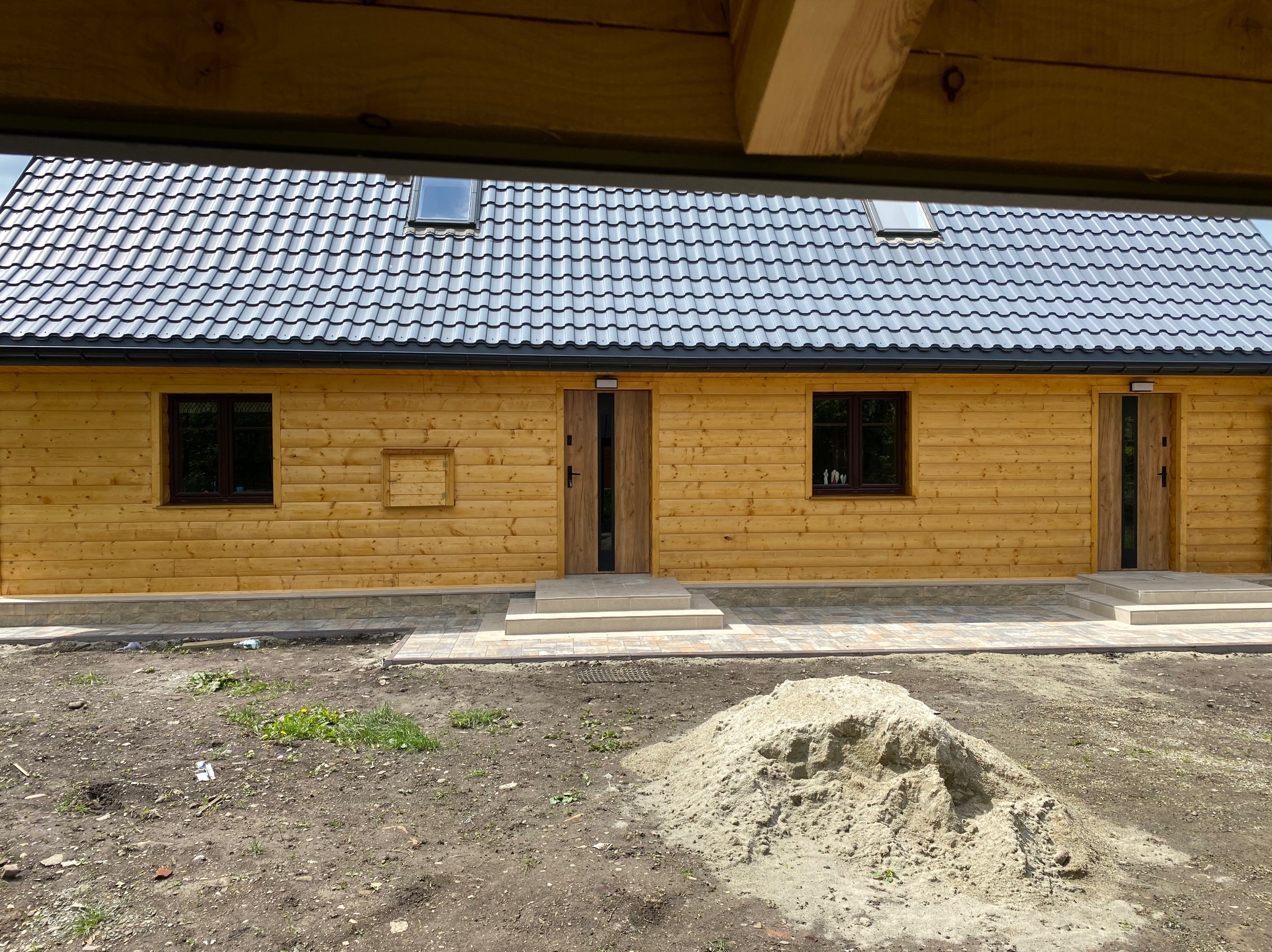 Nowoczesny dom z drewna z szarą dachówką. Widoczne okna dachowe, brązowe okna i drzwi. Na zewnątrz plac budowy z ziemią i piaskiem.