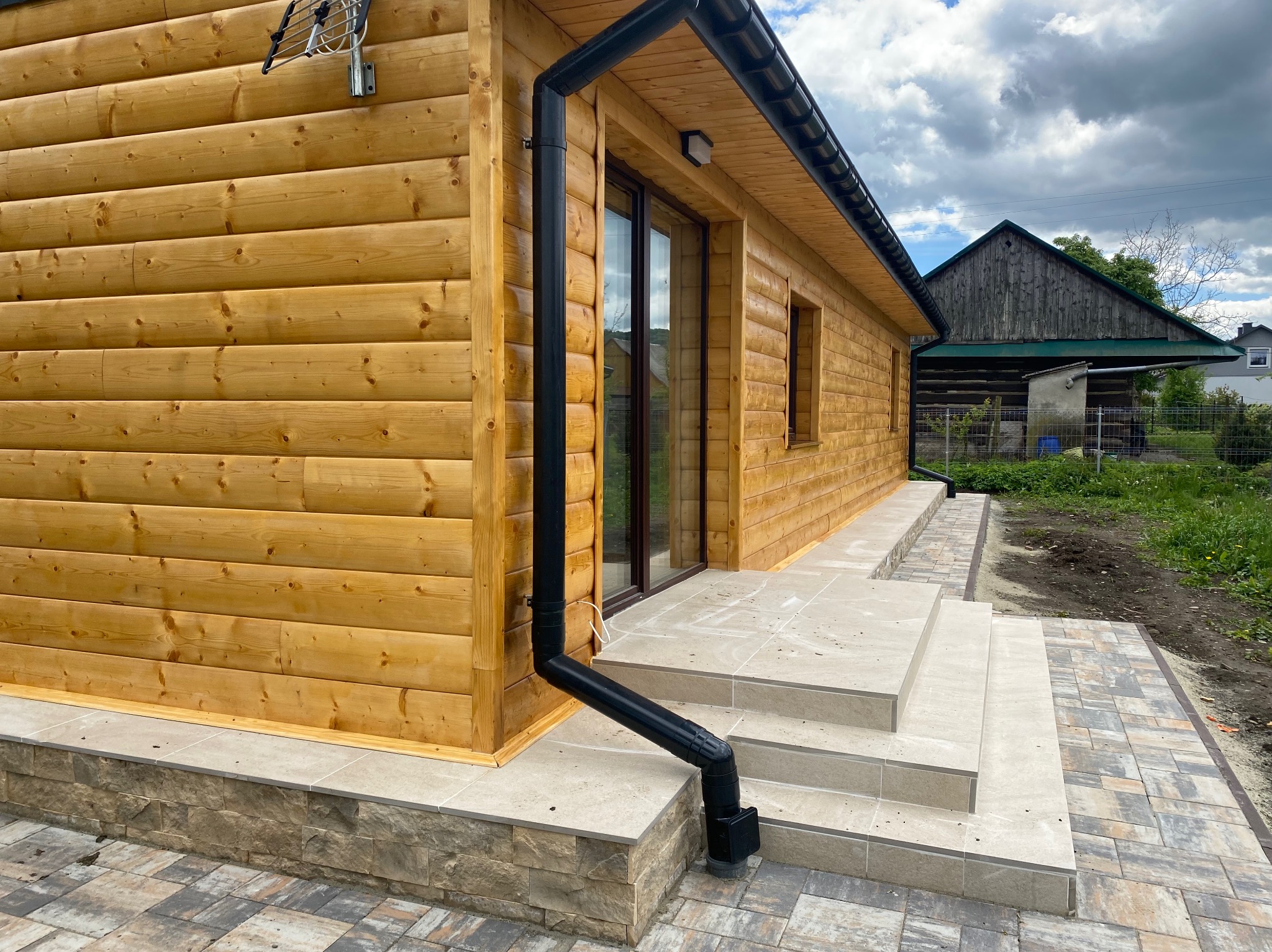 Drewniany dom z czarną rynną i schodami wyłożonymi płytkami. Widoczne detale elewacji i otoczenie budynku w słoneczny dzień.