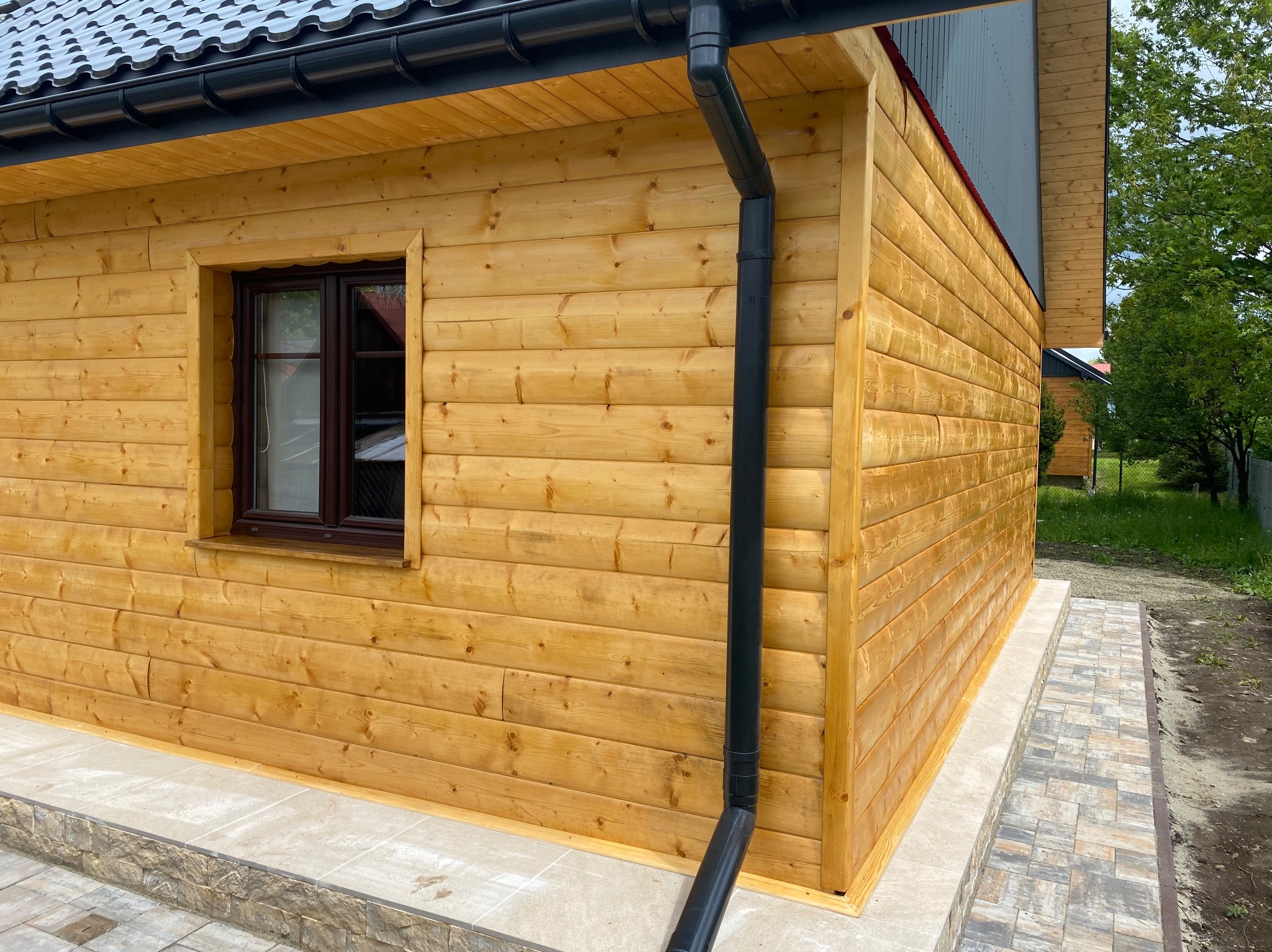 Drewniany domek z brązowym oknem i czarną rynną. Elewacja z jasnego drewna, widoczne łączenia desek. Ujęcie z boku, fragment chodnika z kostki.