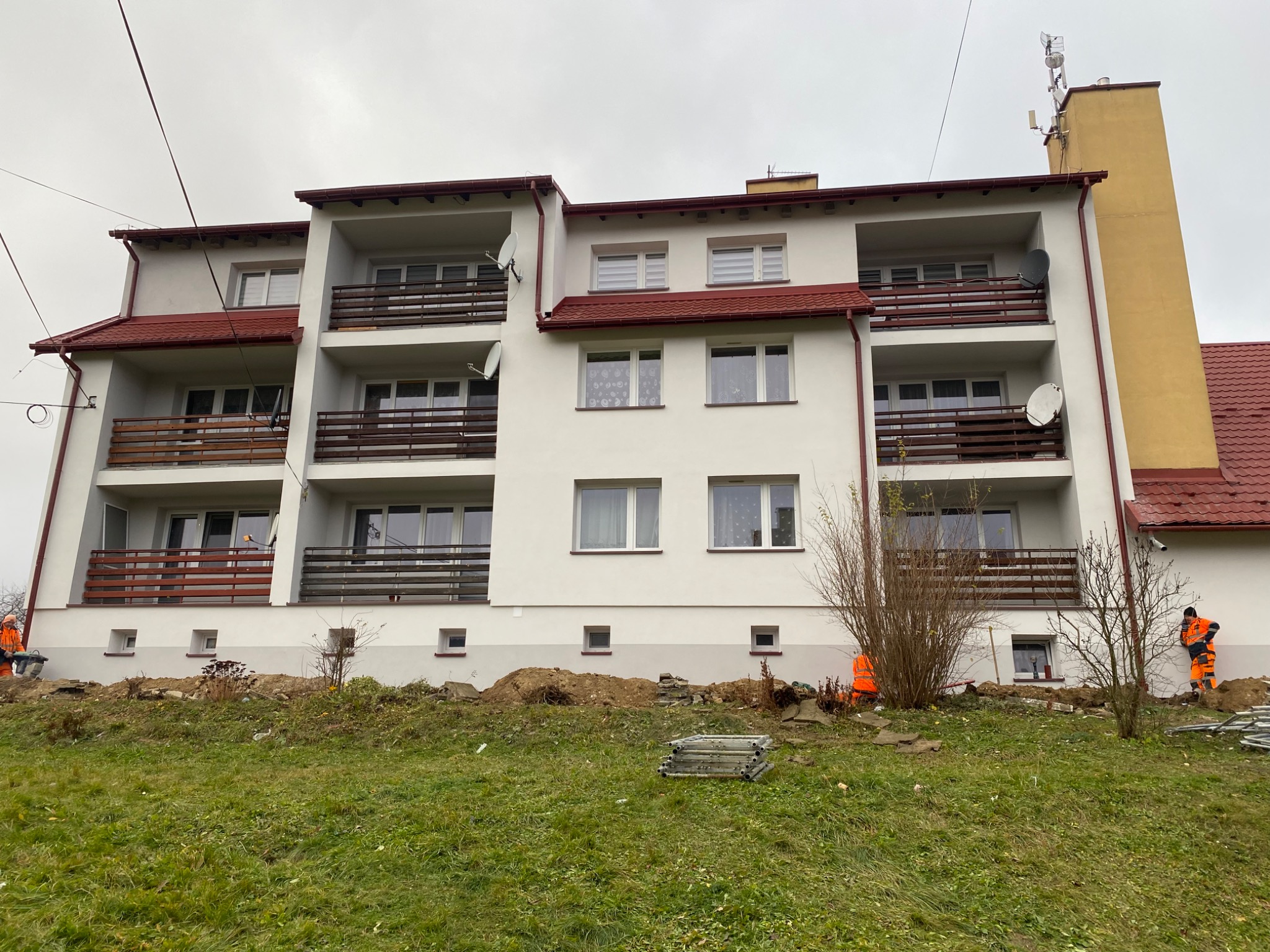 Biały budynek wielorodzinny z balkonami i czerwoną dachówką, widoczni pracownicy w pomarańczowych kombinezonach, teren w trakcie prac ziemnych.