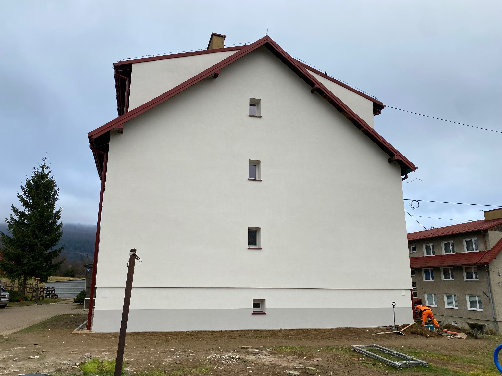 Boczny widok otynkowanego budynku z czerwonym dachem i czterema małymi oknami. Na pierwszym planie pracownik w pomarańczowym kombinezonie.