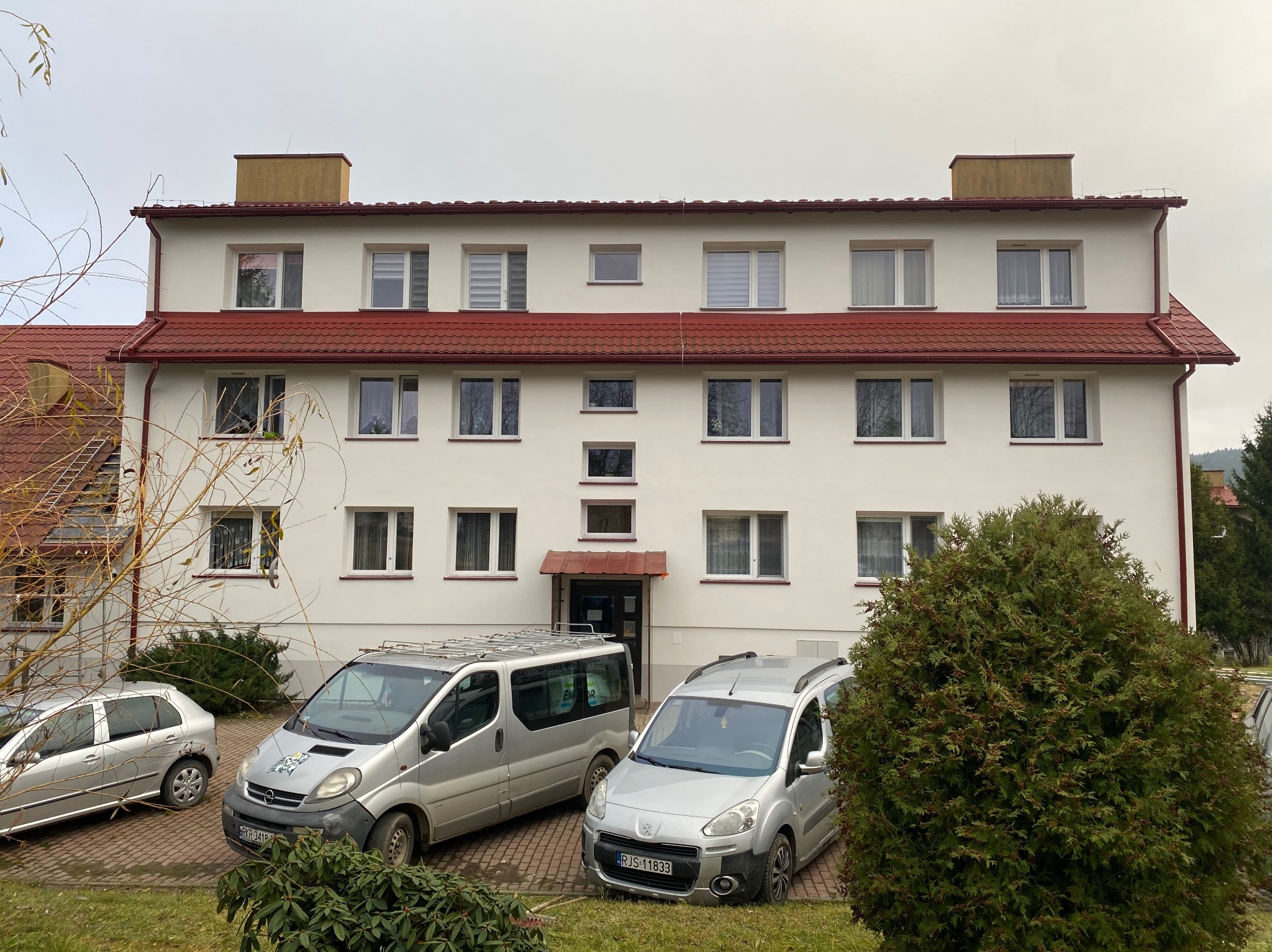 Trzypiętrowy budynek z białą elewacją i czerwonym dachem, widziany z poziomu gruntu. Dwa samochody zaparkowane przed budynkiem. Pochmurne niebo w tle.