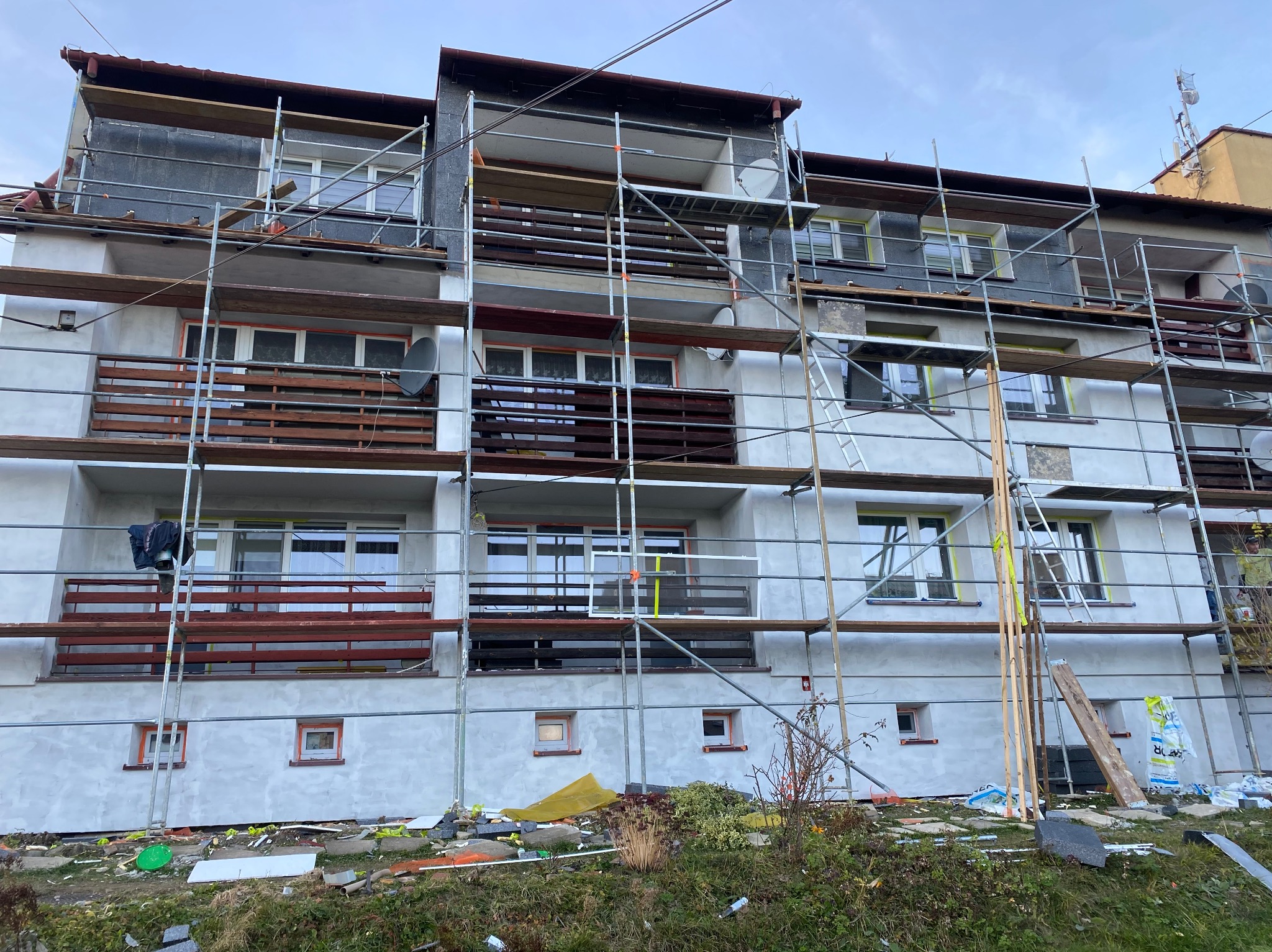 Wielopiętrowy budynek w trakcie ocieplania elewacji, otoczony rusztowaniami. Widoczne fragmenty ocieplonej ściany i przygotowane materiały budowlane.