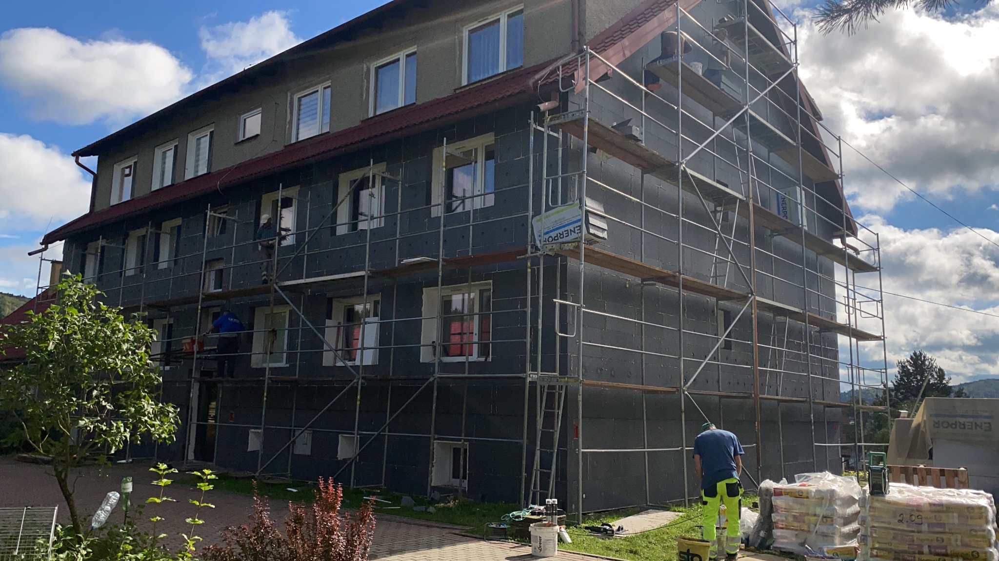 Wielopiętrowy budynek w trakcie ocieplania, pokryty szarym styropianem, z widocznymi rusztowaniami i pracownikami. Częściowo ocieplona elewacja, worki z materiałami budowlanymi.