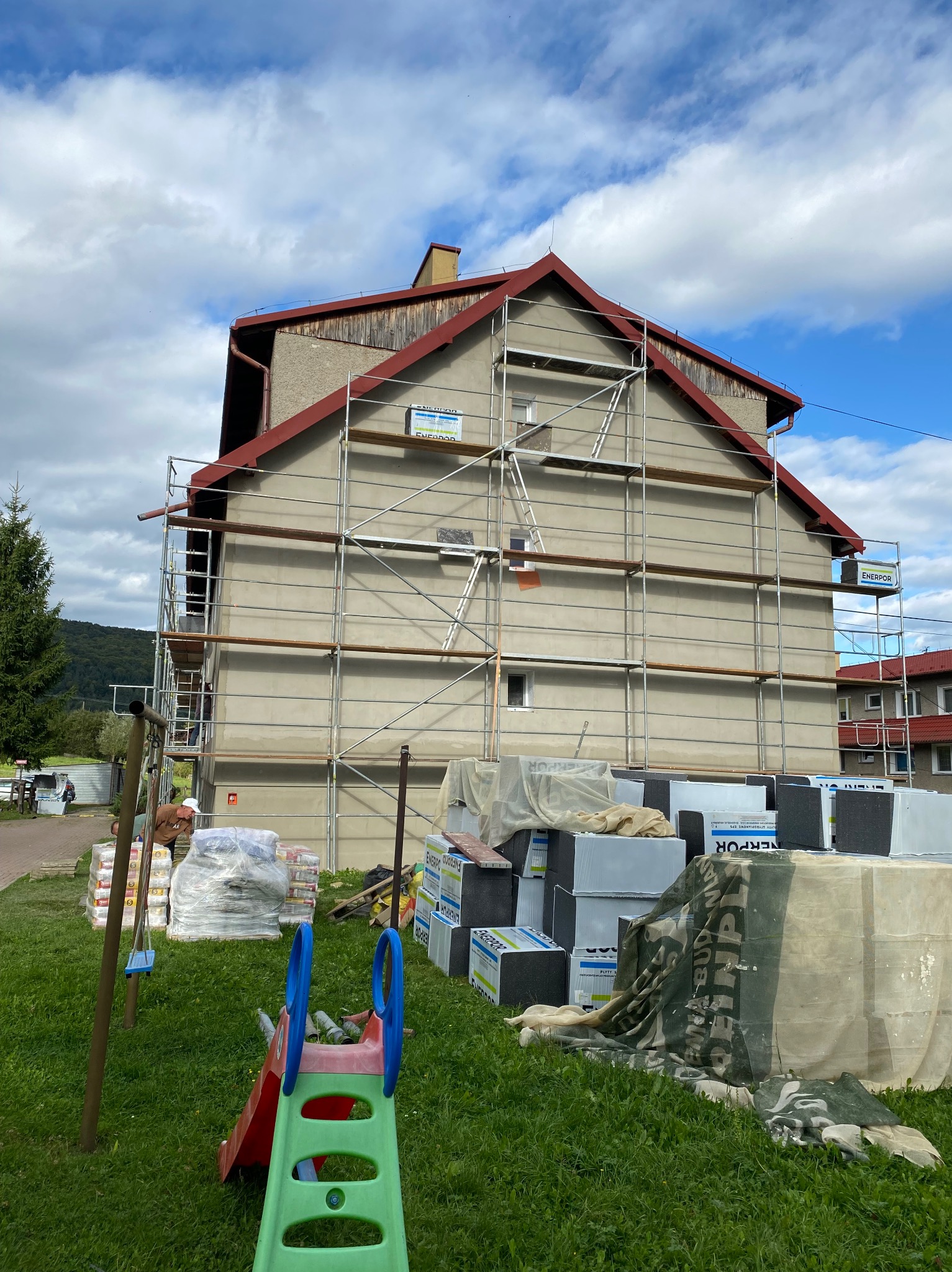 Dom w trakcie ocieplania elewacji, widoczne rusztowania, materiały izolacyjne i częściowo otynkowana ściana. W tle zielony krajobraz i błękitne niebo.