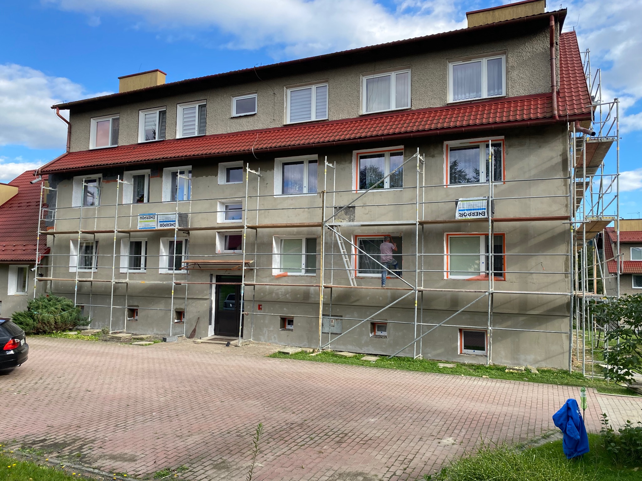 Budynek wielorodzinny w trakcie ocieplania elewacji, widoczne rusztowania i pracownik przy oknie. Częściowo ocieplona ściana z zamontowanymi oknami. Czerwona dachówka.