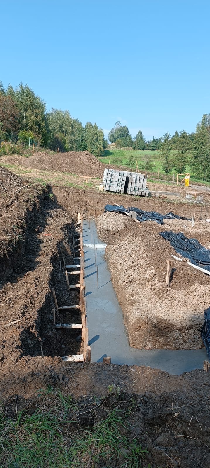 Wylane fundamenty pod budynek, szalunki drewniane, widoczny teren budowy z hałdami ziemi i drzewami w tle.