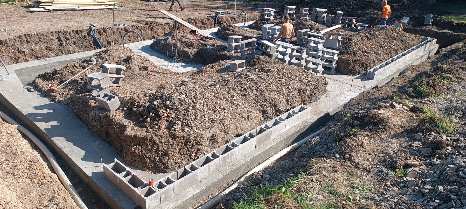 Budowa fundamentów z betonowych bloczków fundamentowych, widoczne zbrojenia i wykop, kilku pracowników na budowie.