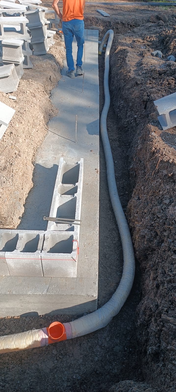 Wykop z betonową wylewką fundamentową, częściowo wymurowany z bloczków betonowych, widoczna rura kanalizacyjna w folii ochronnej i przewód wentylacyjny, na górze widoczna osoba w pomarańczowej...