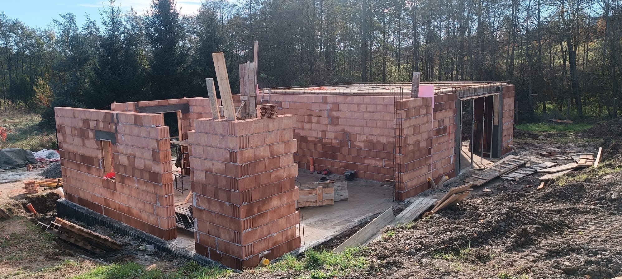 Wznoszenie ścian parteru z czerwonej cegły na budowie domu jednorodzinnego, widoczne zbrojenie i przygotowania do zalania wieńca.