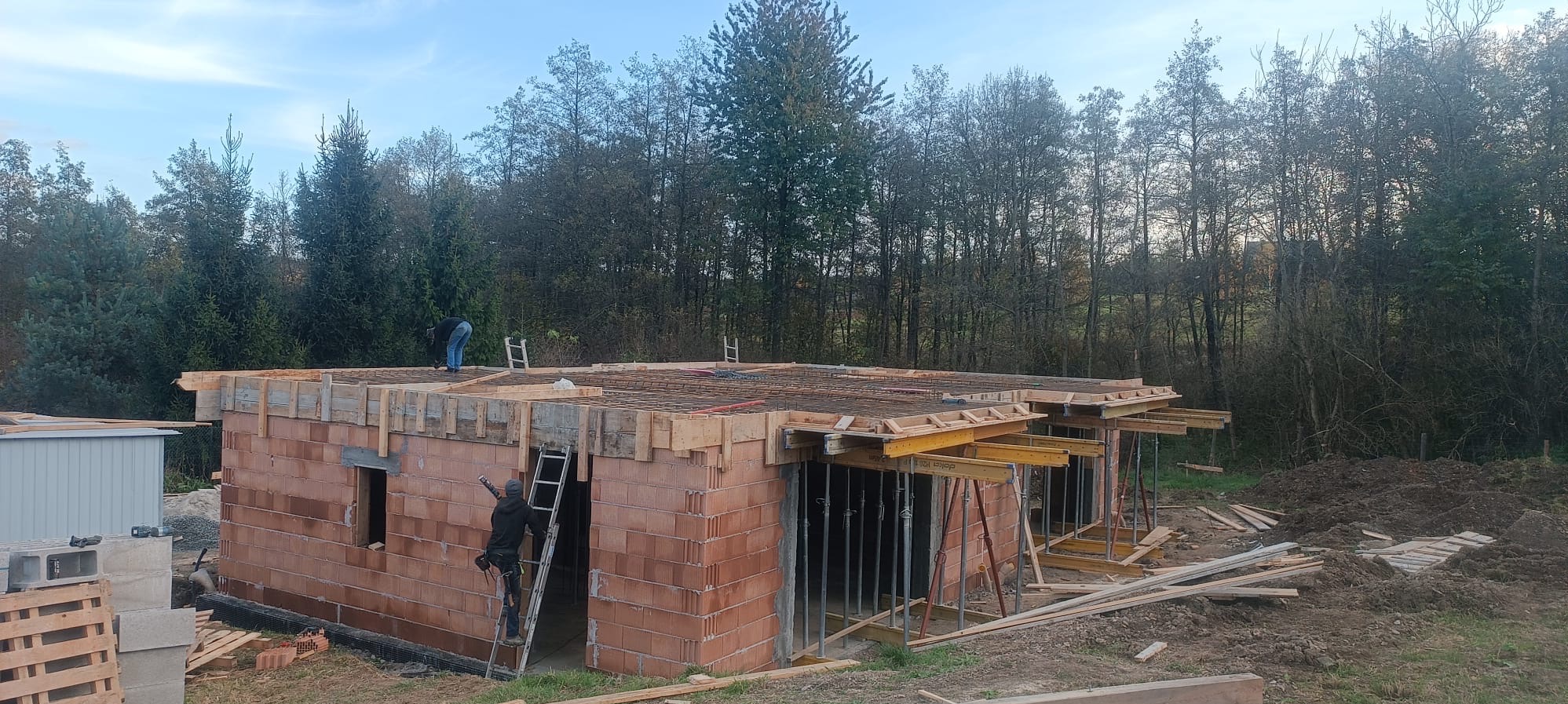 Wznoszenie ścian z czerwonej cegły i zbrojenia stropu w dwukondygnacyjnym budynku w stanie surowym otwartym, widoczne podpory stropowe i pracownicy na budowie.