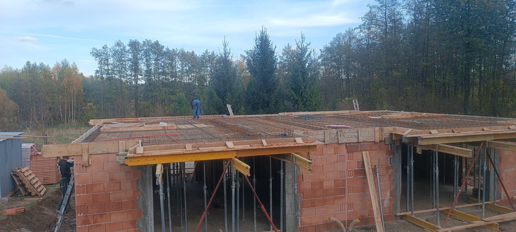 Budowa domu z czerwonej cegły: zbrojona konstrukcja stropu z deskowaniem i podporami, pracownik na górze, widok na las w tle.