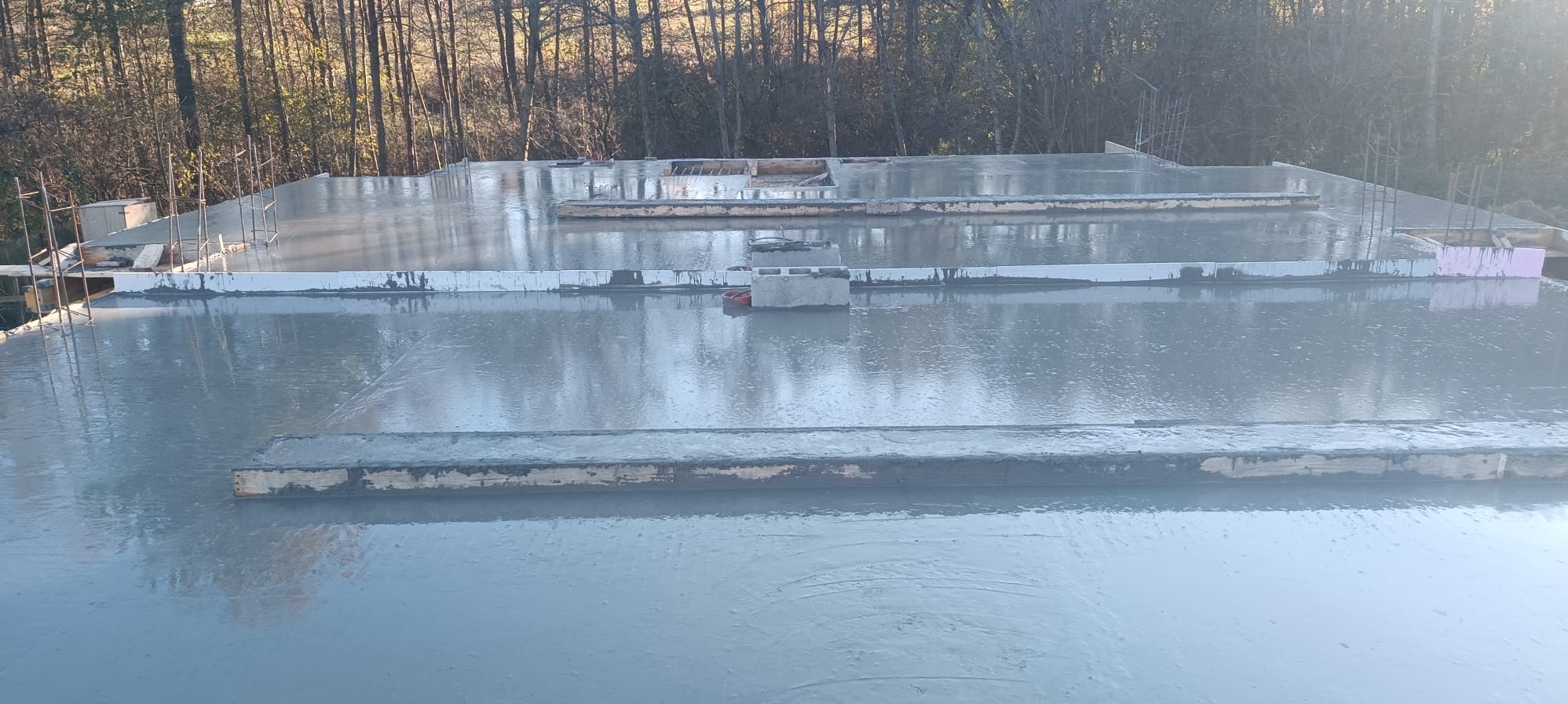 Wylany, mokry beton na przygotowanym fundamencie pod budowę, widoczne zbrojenia i szalunki, w tle drzewa.