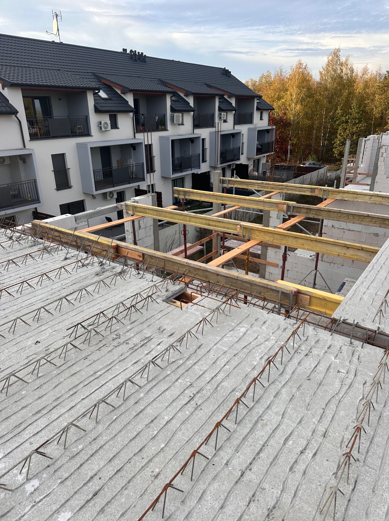 Wiązanie zbrojenia stropu monolitycznego na budowie, widoczne podpory szalunkowe i fragmenty ścian z betonu komórkowego, w tle budynek mieszkalny z balkonami i drzewa z jesiennymi liśćmi.
