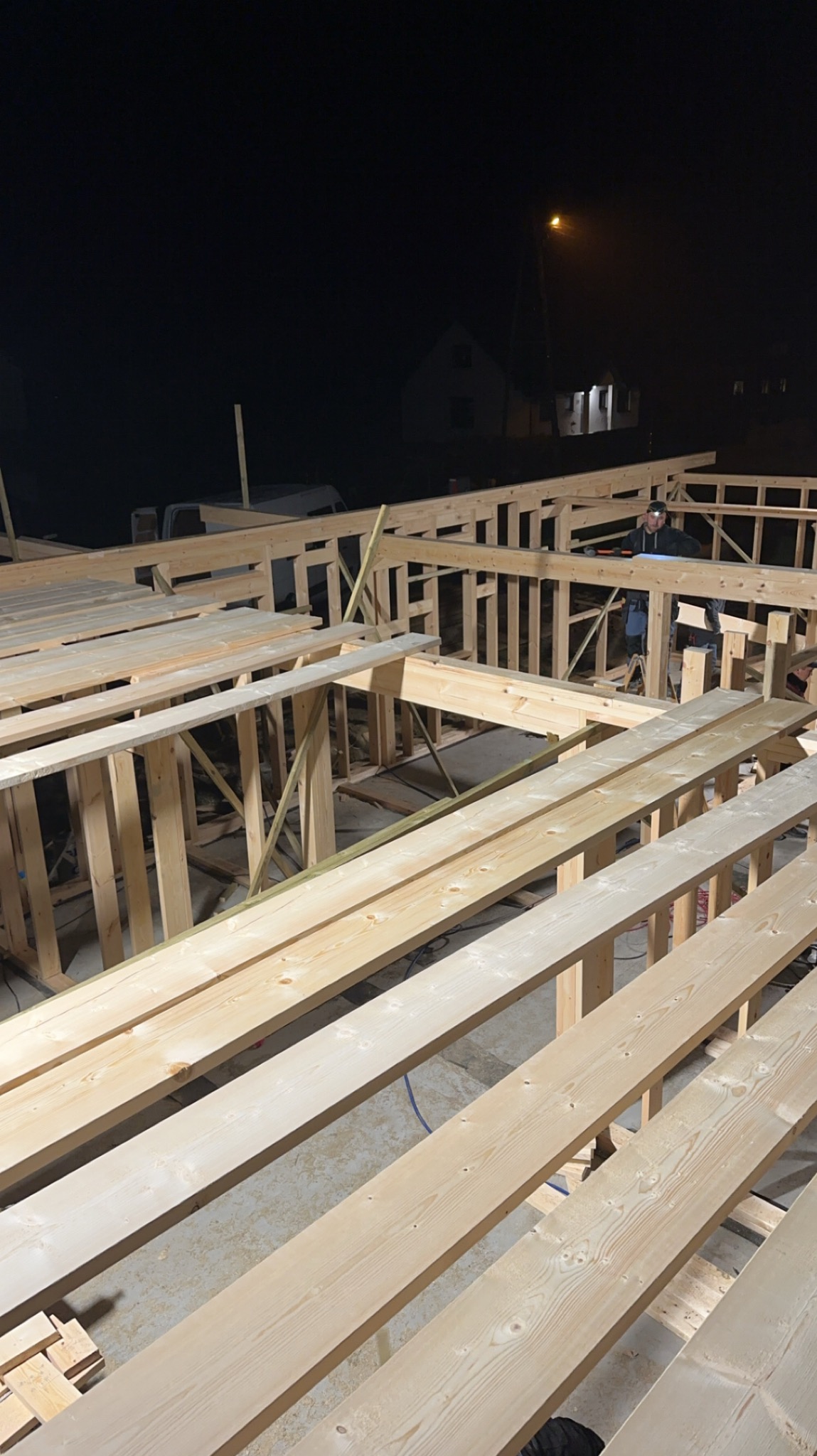 Konstrukcja szkieletowa domu w trakcie montażu nocą, widoczne drewniane belki i słupy, w tle oświetlony budynek i lampa uliczna, pracownik w odzieży roboczej z narzędziami.